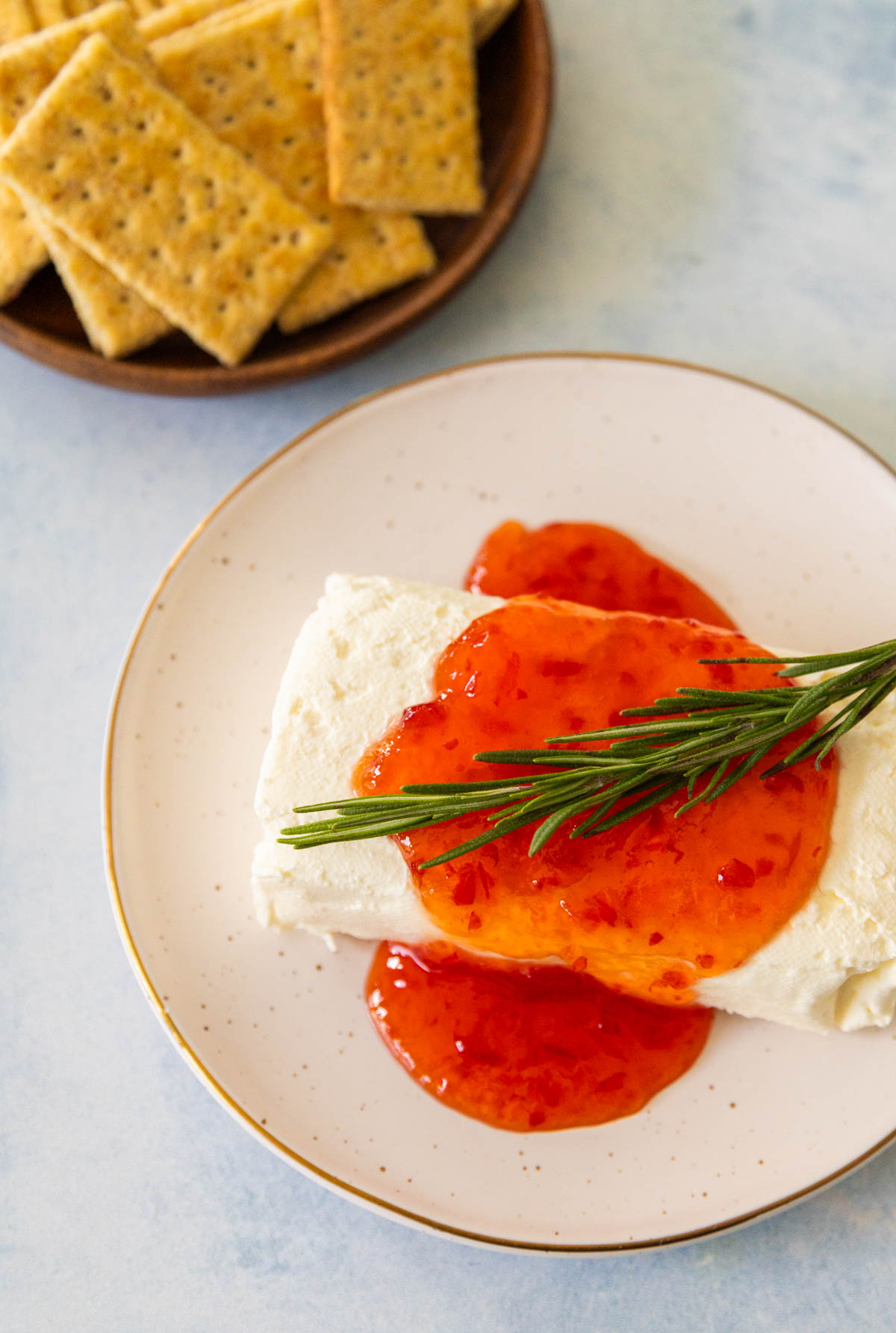 A block of cream cheese on a plate has pepper jelly spooned over the top with a sprig of fresh rosemary. A plate of crackers is in the background.