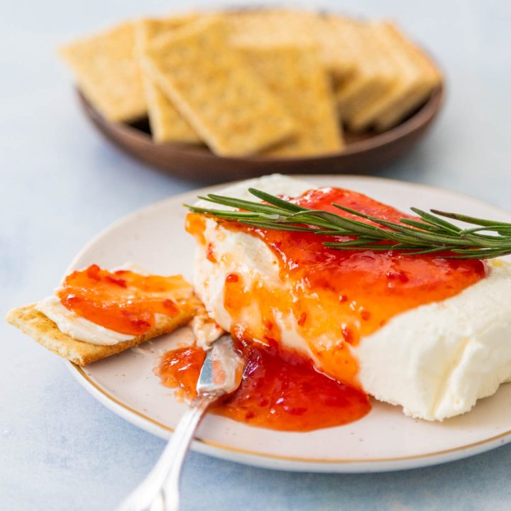 A block of cream cheese on a plate has pepper jelly spooned over the top with a sprig of fresh rosemary. A plate of crackers is in the background.