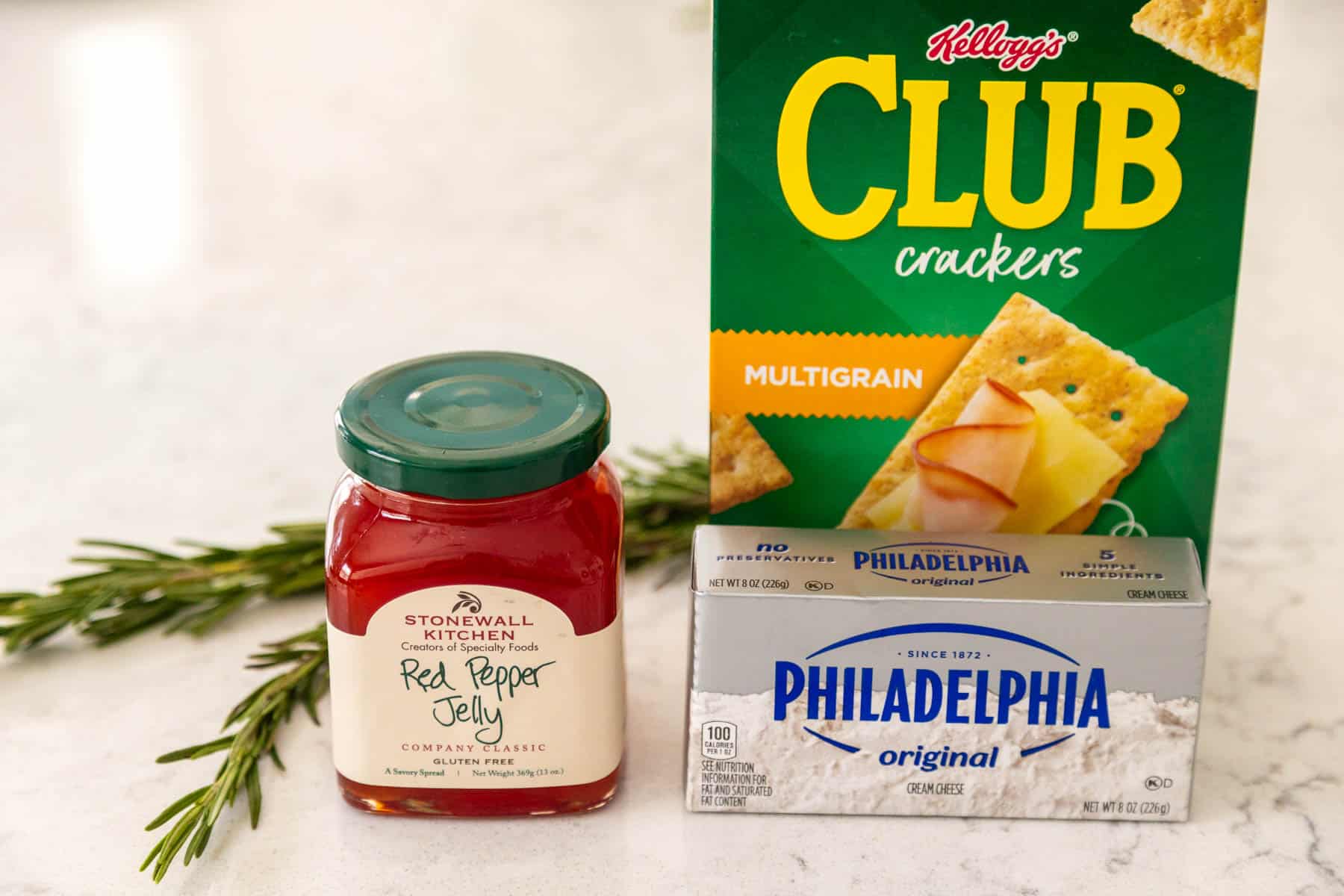 A jar of pepper jelly sits next to a brick of cream cheese and a box of crackers. A fresh sprig of rosemary sits behind.