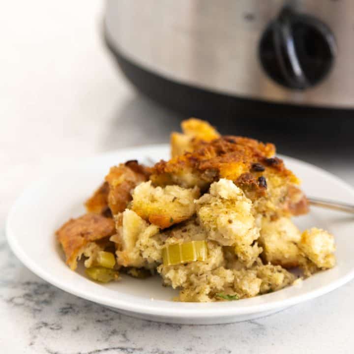 A scoop of golden brown Crock Pot stuffing is served on a small white plate. The slowcooker power knob is seen in the back.