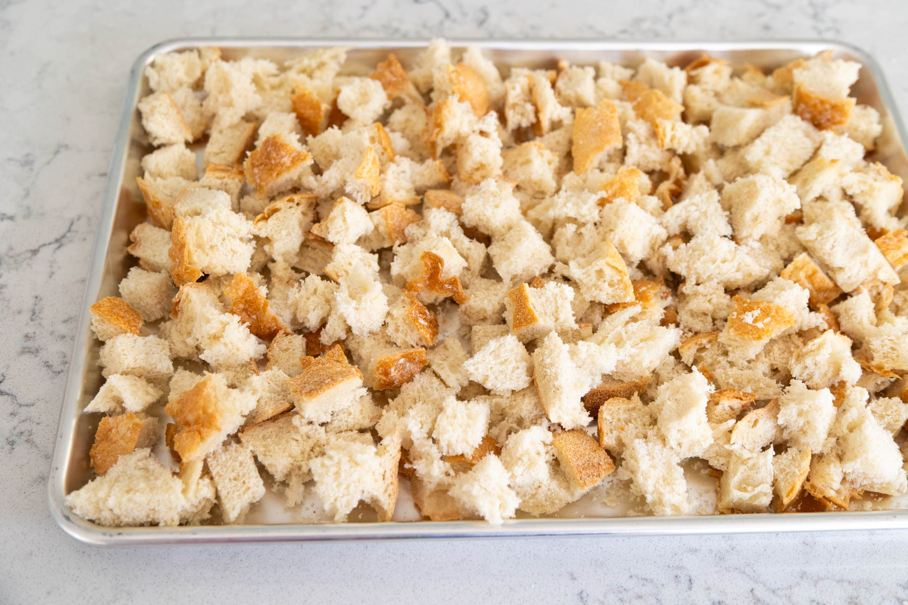 A large metal baking sheet is filled with hand ripped bread chunks to make homemade stuffing croutons in the oven.