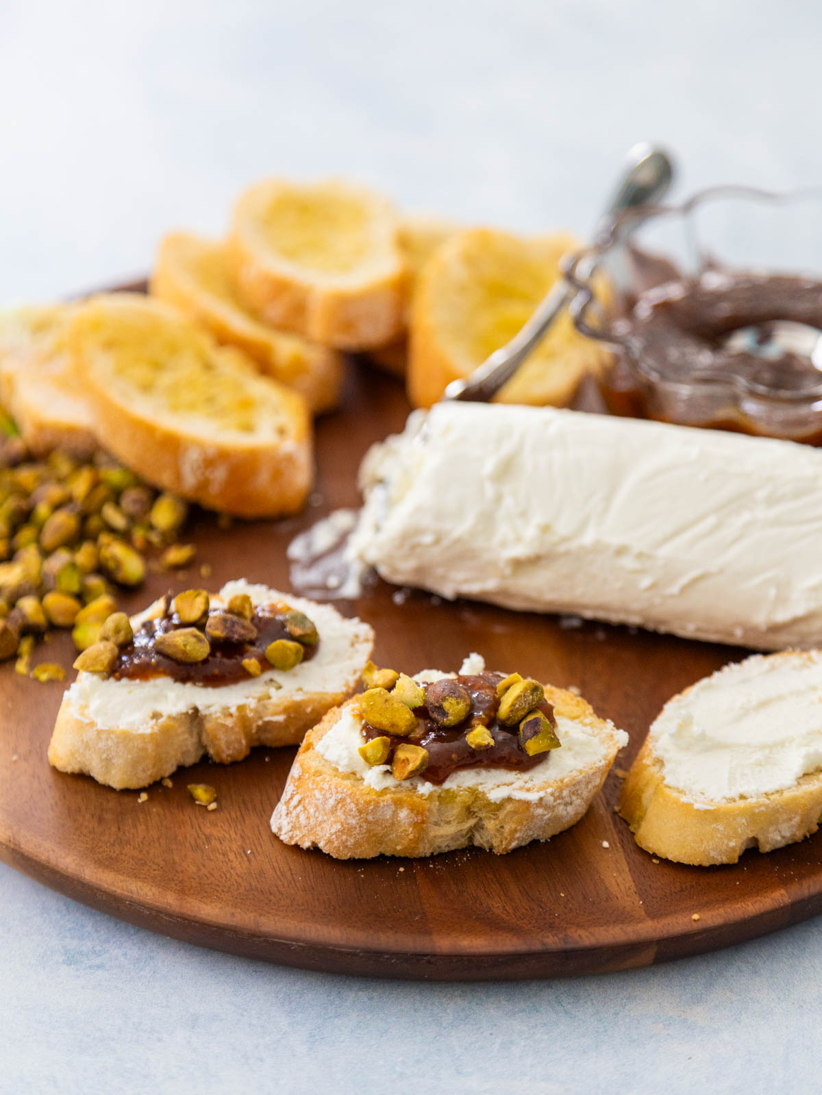 A charcuterie platter has crostini, soft goat cheese, chopped pistachios, and a bowl of fig jam. A few assembled crostini bites are in front.