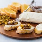 A charcuterie platter has crostini, soft goat cheese, chopped pistachios, and a bowl of fig jam. A few assembled crostini bites are in front.