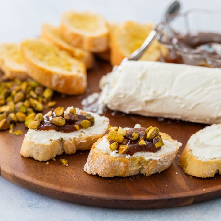 A charcuterie platter has crostini, soft goat cheese, chopped pistachios, and a bowl of fig jam. A few assembled crostini bites are in front.