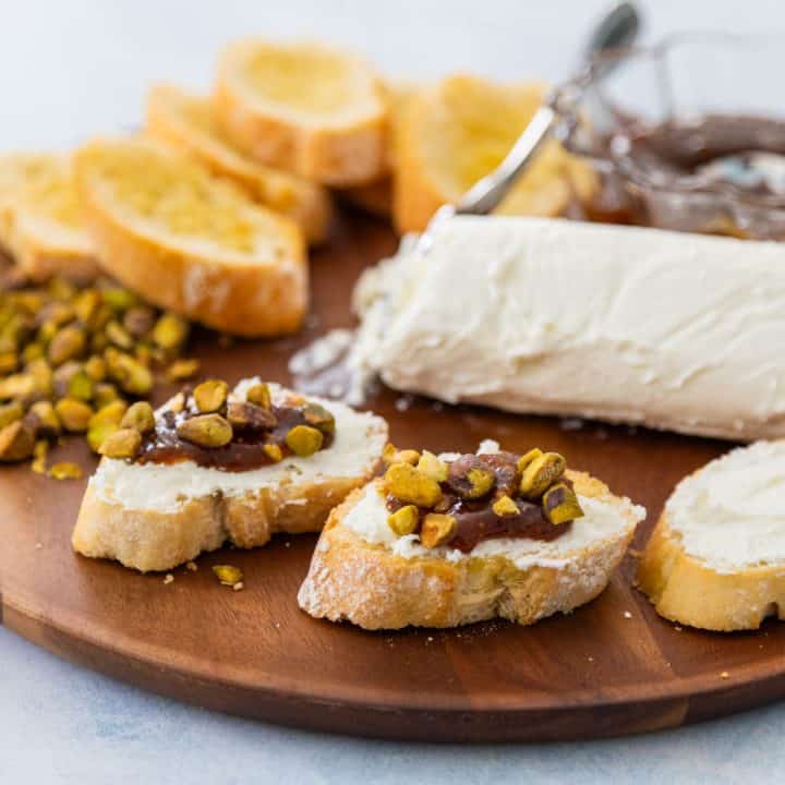 A charcuterie platter has crostini, soft goat cheese, chopped pistachios, and a bowl of fig jam. A few assembled crostini bites are in front.