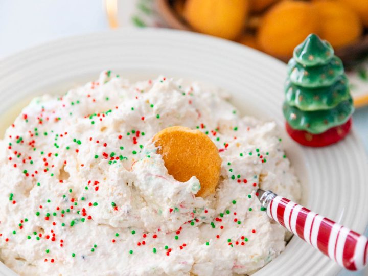A bowl filled with a light and fluffy white dessert dip with red and green sprinkles has a wafer cookie dunked in it and a peppermint striped spoon stirring it.