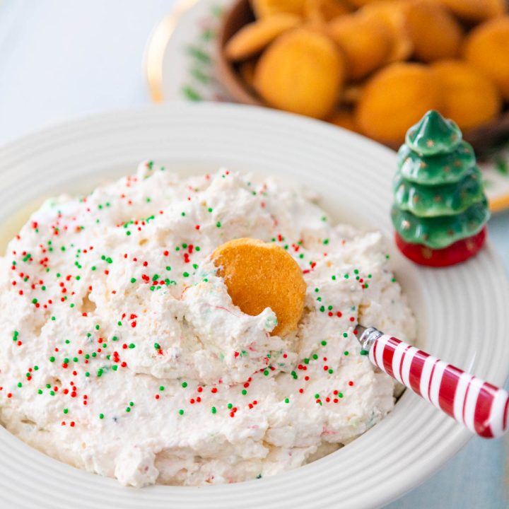 A bowl filled with a light and fluffy white dessert dip with red and green sprinkles has a wafer cookie dunked in it and a peppermint striped spoon stirring it.