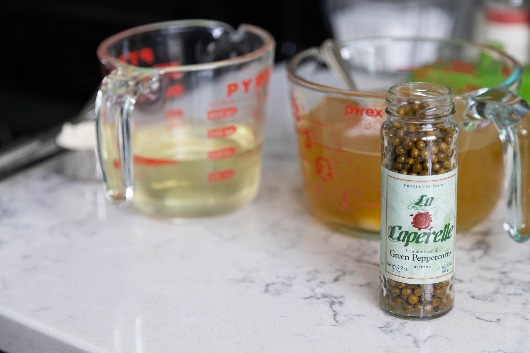 The jar of green peppercorns is near the measuring cup of wine and chicken stock.
