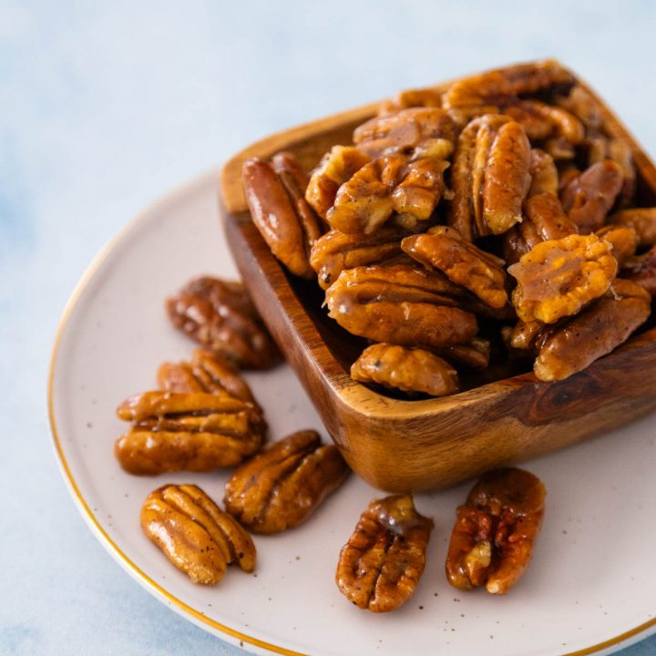A wooden bowl on a plate is overflowing with glazed pecans.