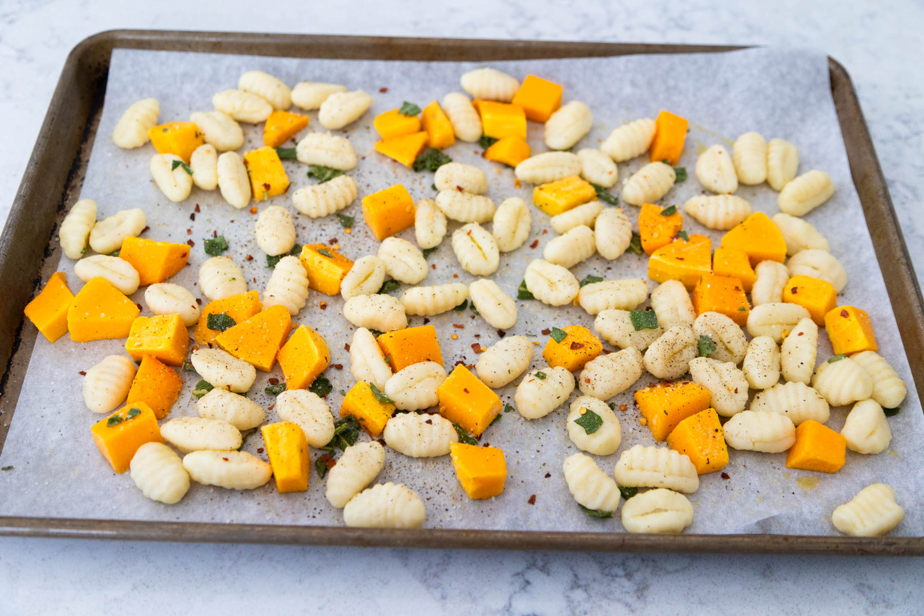 A baking pan lined with parchment paper has the gnocchi and butternut squash spread out into an even layer.