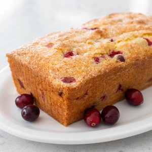 A loaf of cranberry bread is on a white platter with fresh cranberries sprinkled around for garnish.
