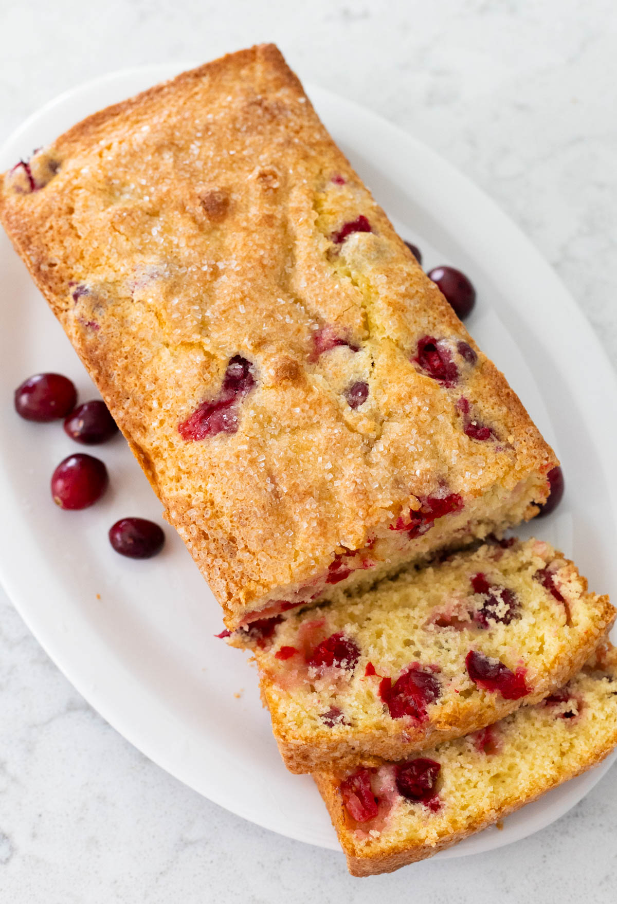 The loaf of homemade cranberry bread is on a white platter, two slices have been sliced so you can see the texture of the quick bread and the large, whole cranberries.