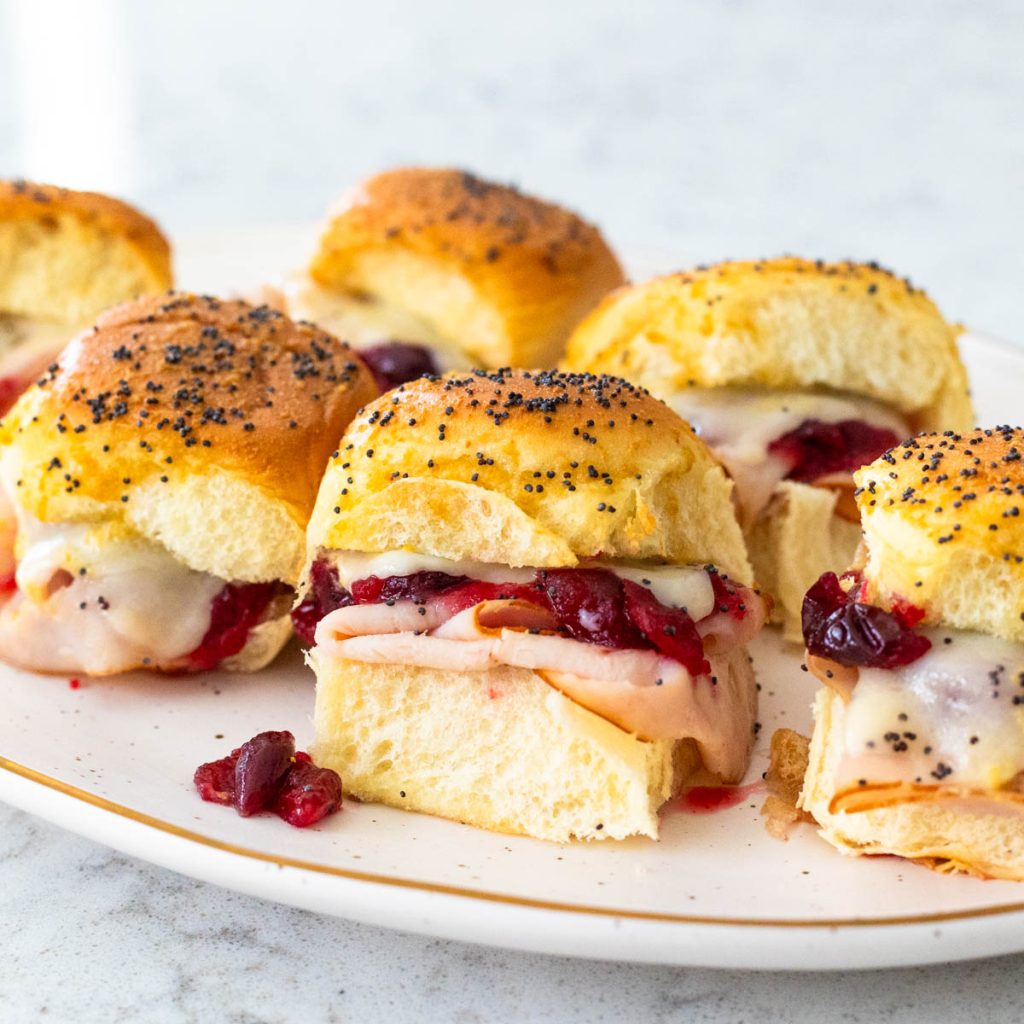 A platter of cranberry turkey sliders show the melted cheese and poppy seeds on top.