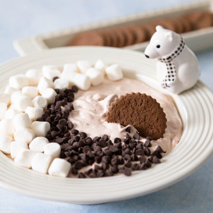 A white bowl filled with chocolate dip that has marshmallows and chocolate chips on top. A chocolate cookie is being dunked into the dip.