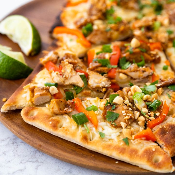 The baked Thai chicken pizza has chopped peanuts, green onions, and red pepper strips on top. Two lime wedges are on the side of the plate.