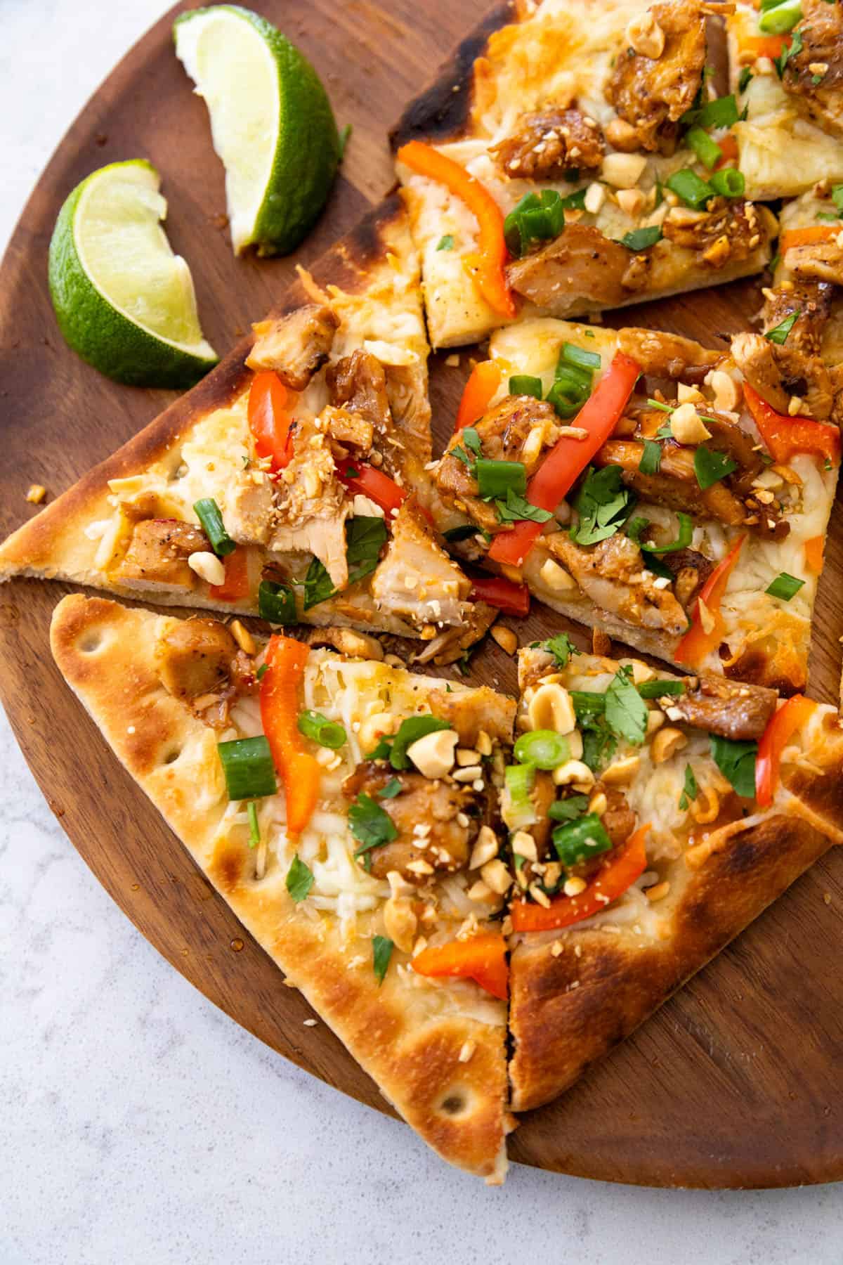 The baked Thai chicken pizza has chopped peanuts, green onions, and red pepper strips on top. Two lime wedges are on the side of the plate.