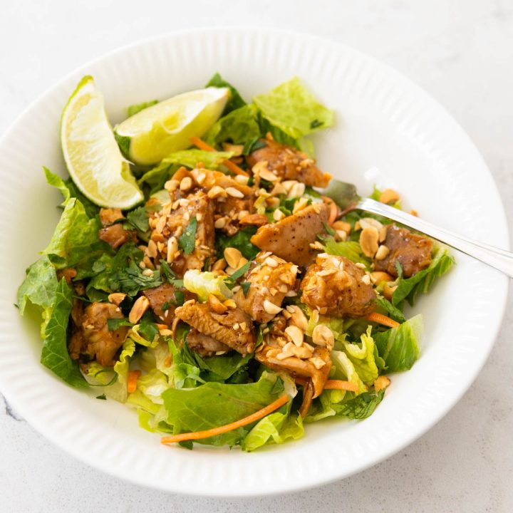 A white bowl is filled with a light green salad topped with Thai chicken and lime wedges.