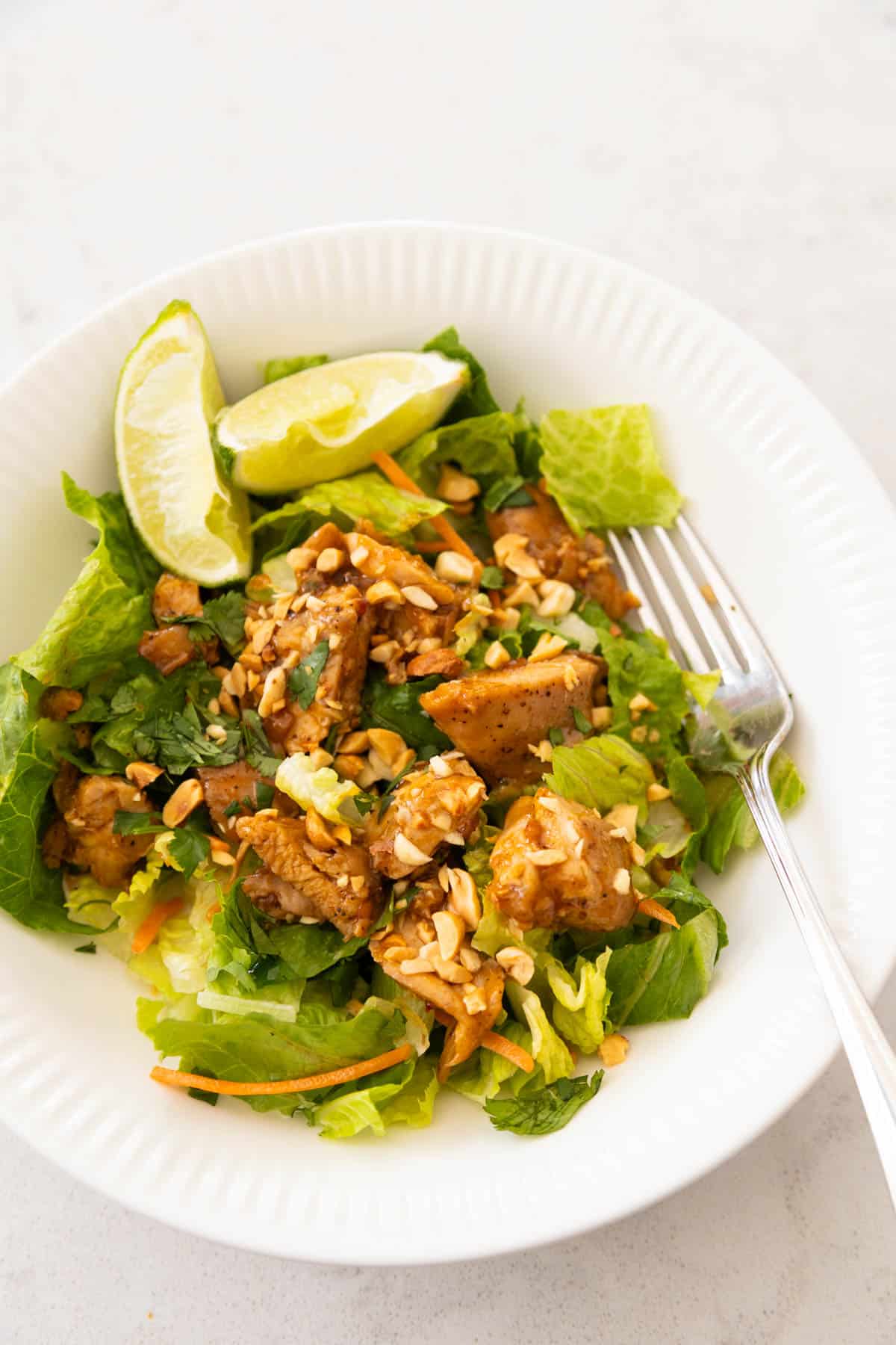 A white bowl is filled with a light green salad topped with Thai chicken and lime wedges.