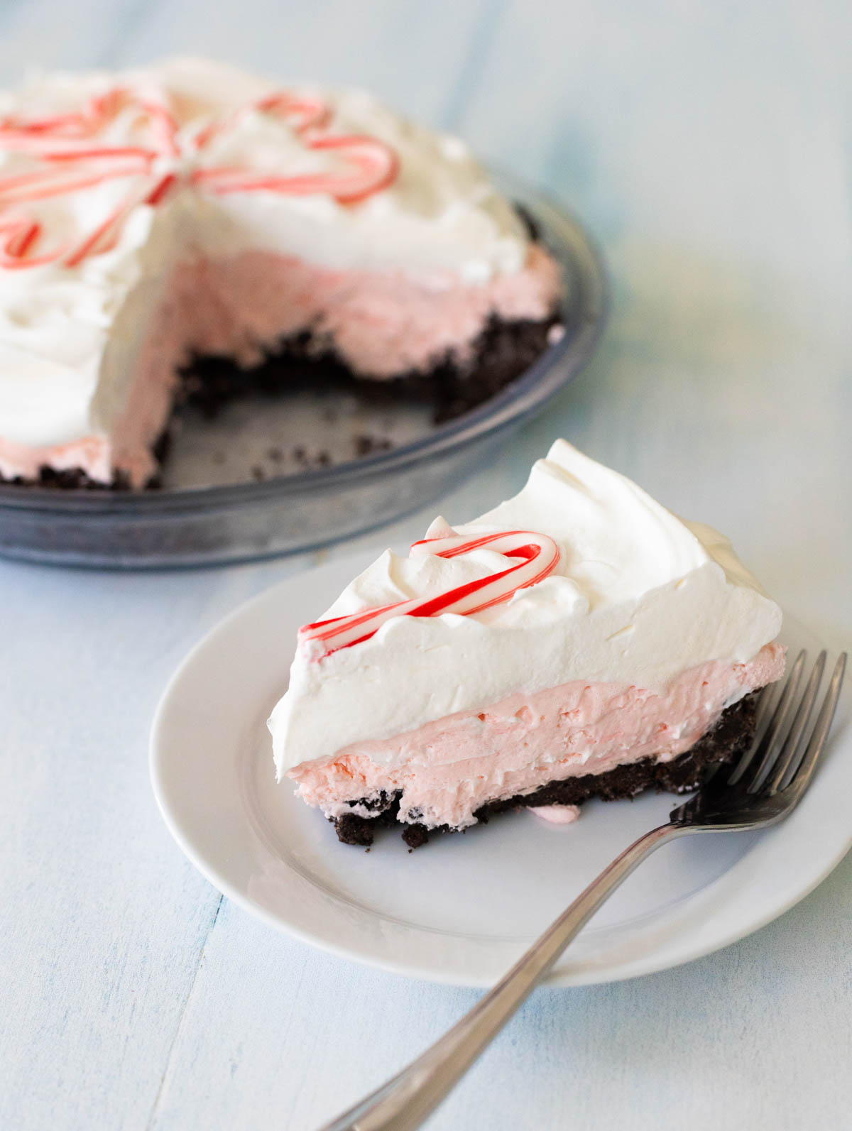 A slice of the candy cane pie is on a white plate with a fork so you can see the layers of creamy filling. The sliced pie is in the background.