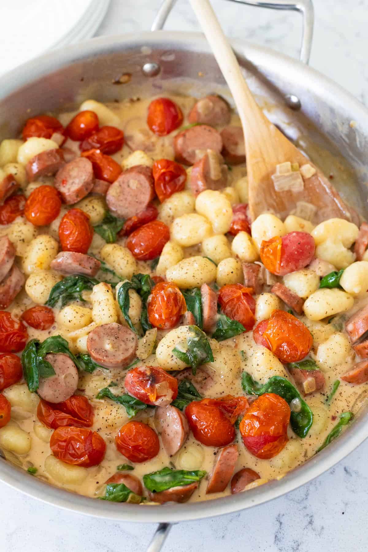 A skillet of creamy gnocchi is being stirred with a wooden spoon and about to be served.