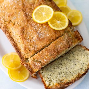 A loaf of lemon poppy seed bread is on a platter, a few pieces have been sliced. Sliced lemons are added as garnish.