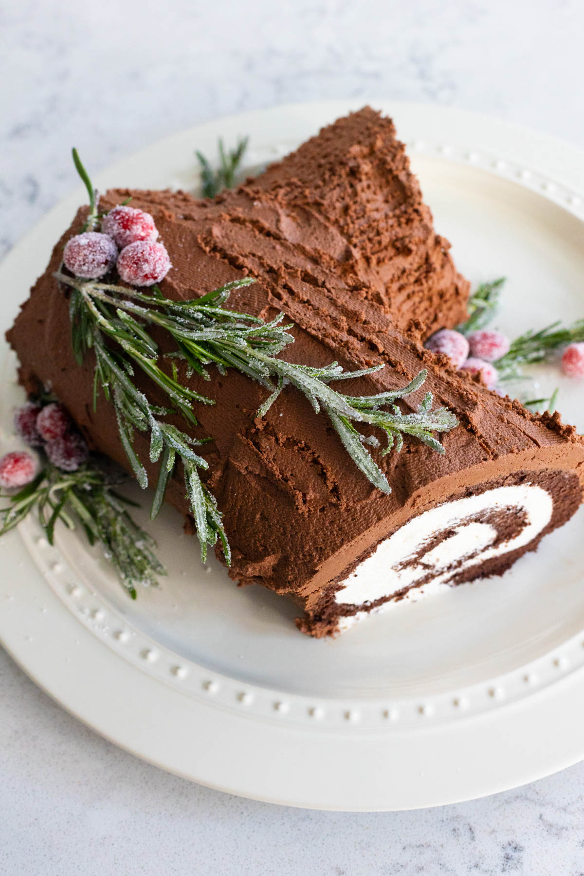 The finished yule log cake sits on a cake plate with sugared cranberries and rosemary.