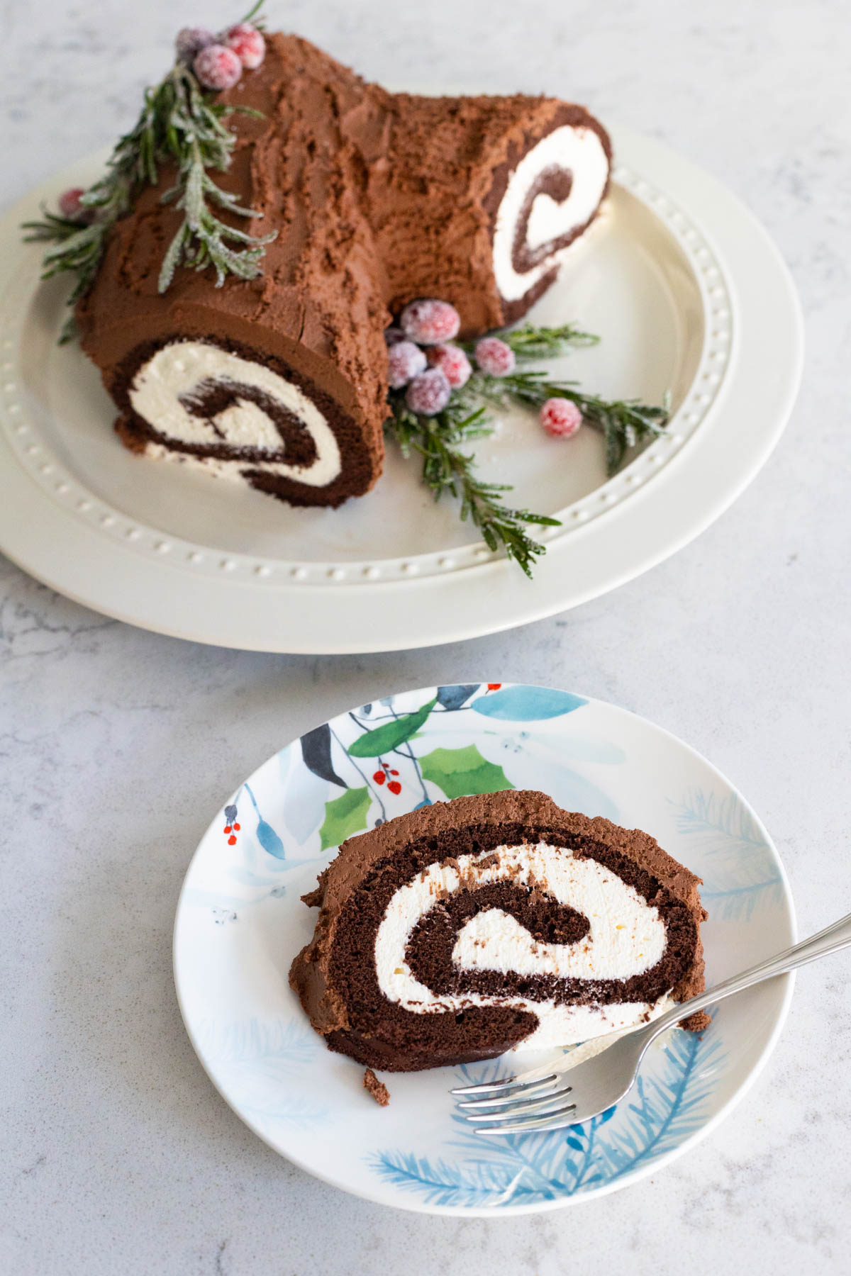 The sliced yule log cake is on a Christmas plate with a fork. The whole yule log cake is on the platter in the background.