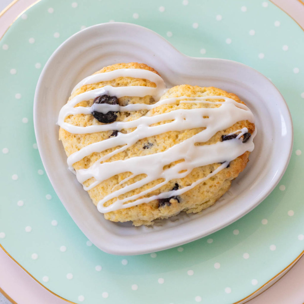 A heart-shaped almond cherry scone with a drizzle of icing over the top is in a heart-shaped plate.