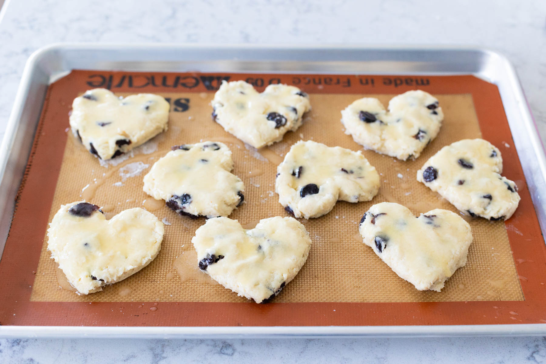The scones have been cut into heart shapes and brushed with melted butter on a baking sheet lined with a silicone baking mat.
