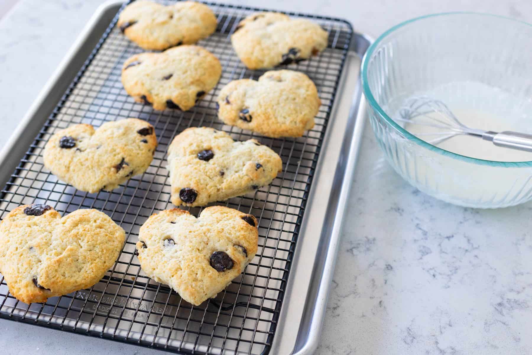 The scones are on a wire rack next to a bowl of almond icing with a whisk.