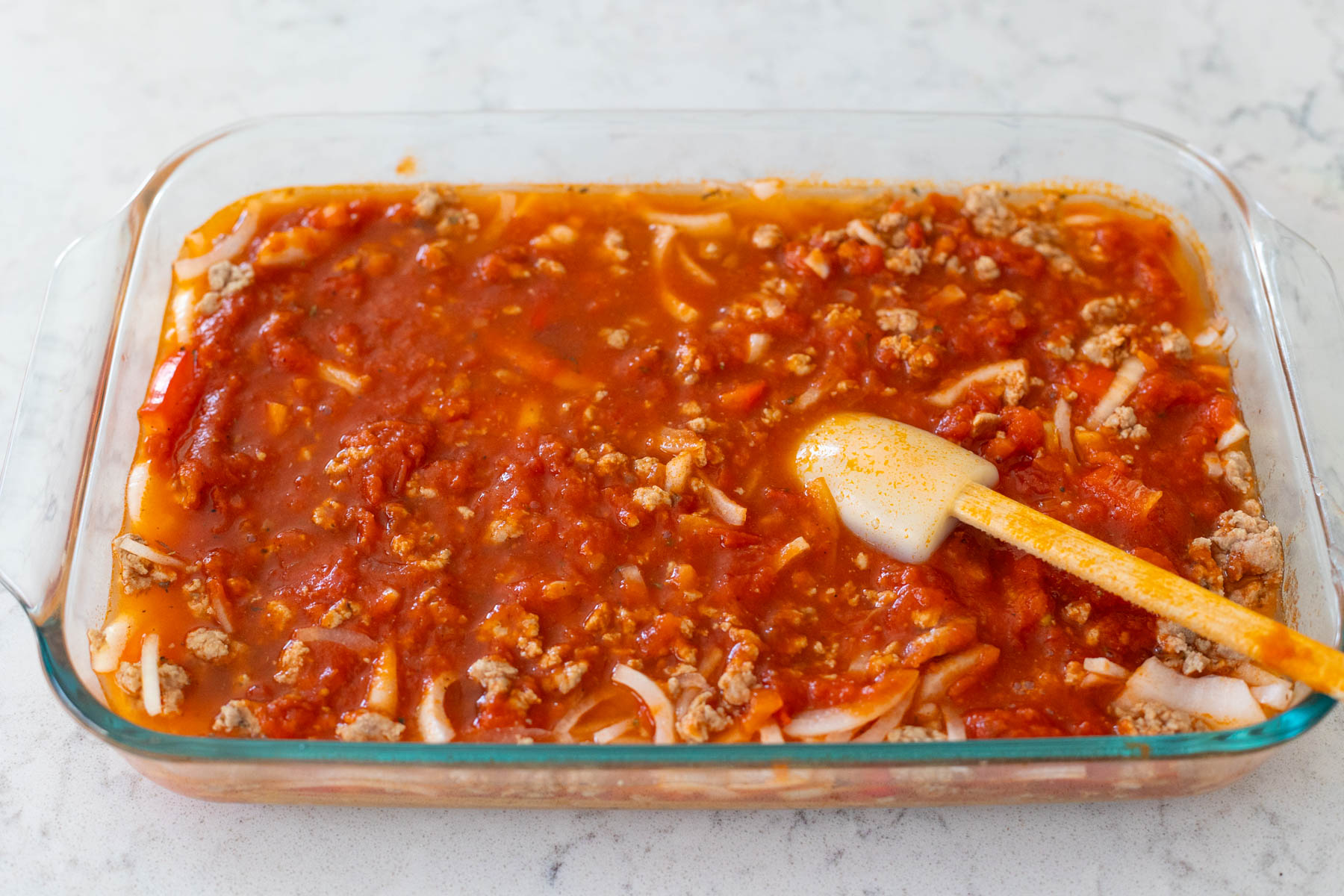 The spatula has just finished gently mixing the water and tomato sauce together in the baking dish.