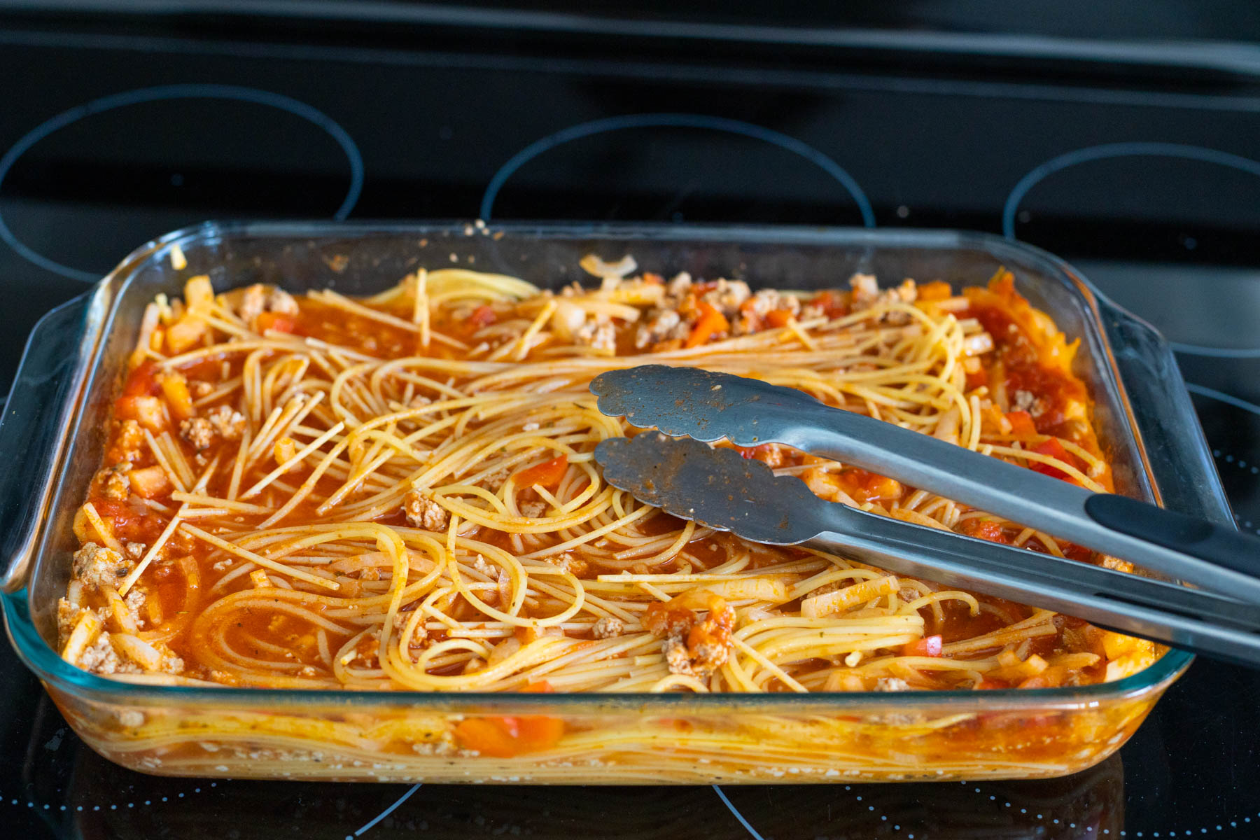 The tongs have just finished turning the spaghetti in the baking dish.