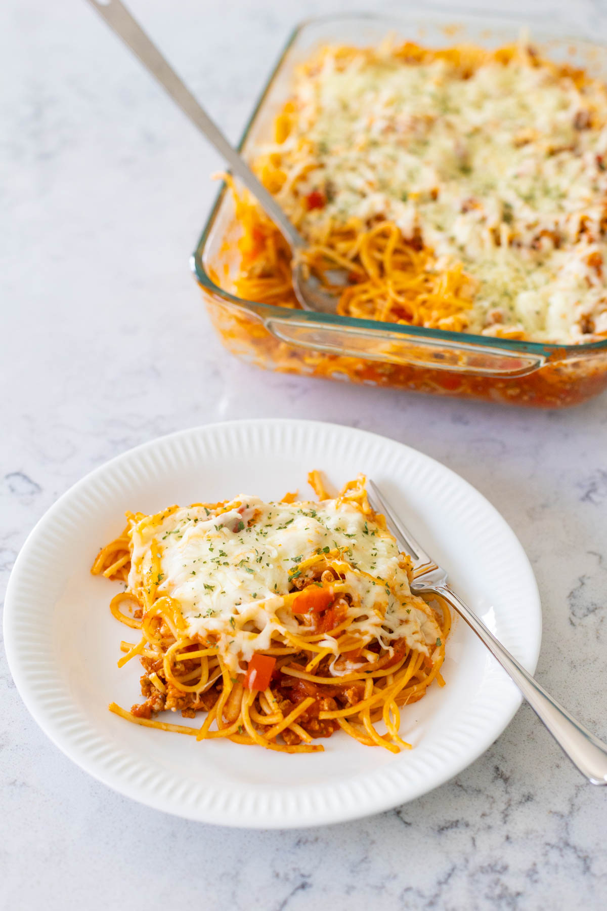 A portion of baked spaghetti has been served up onto a white plate with a fork. The baking dish is in the background with a serving spoon sticking out of it.