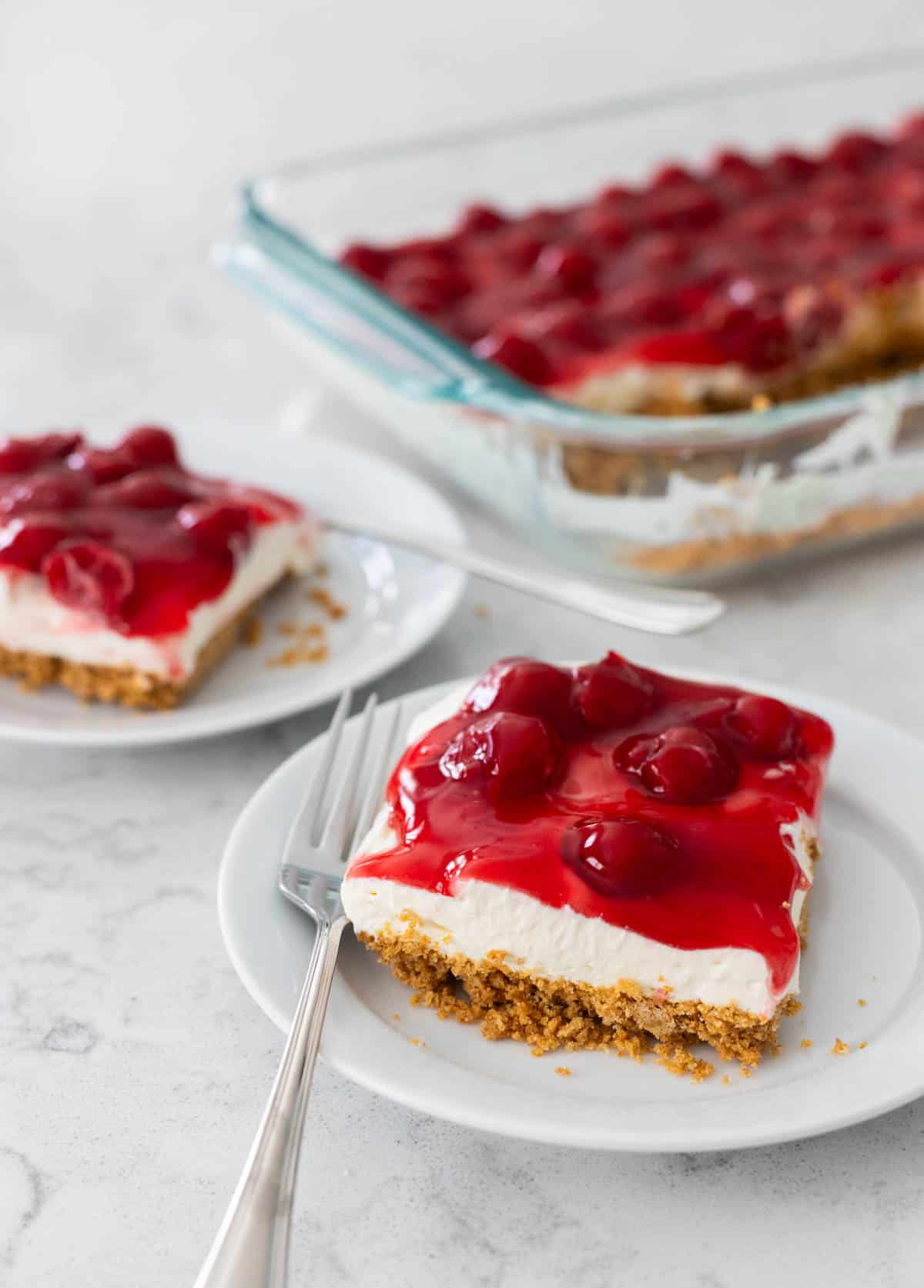 Two servings of cherry delight are on white plates next to the baking pan that has the rest of the dessert.