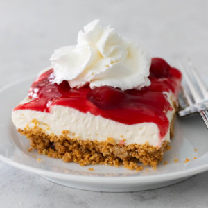 A layered cherry delight dessert slice on a white plate. You can see layers of graham cracker crust, cream cheese filling, and cherry pie filling on top with a dollop of whipped cream.
