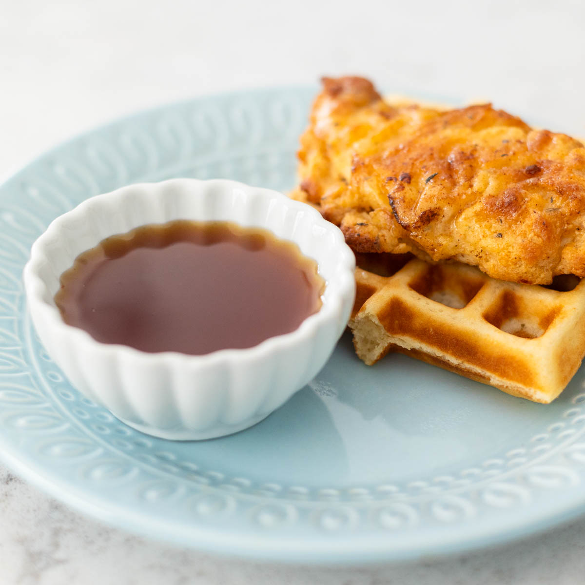 A cup of sauce sits on a plate next to a fried chicken tender on a waffle.