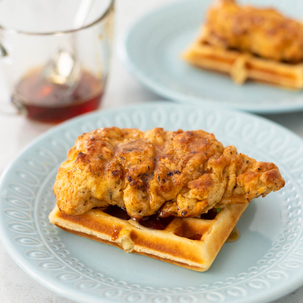 A buttermilk waffle has buttermilk fried chicken on top and a special pitcher of spiced maple syrup in the back.