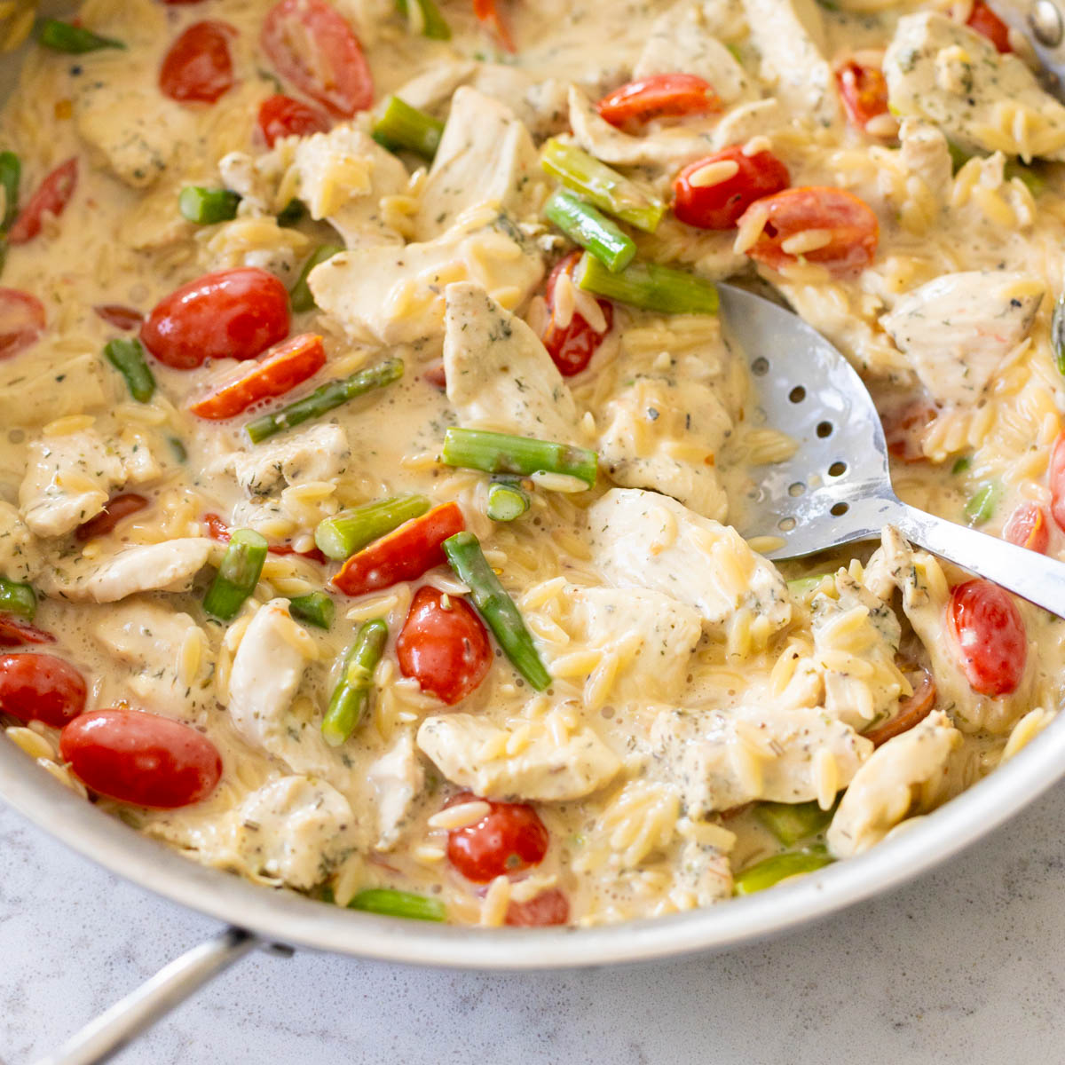 A skillet is filled with creamy orzo with chicken chunks, asparagus spears, and cherry tomatoes.