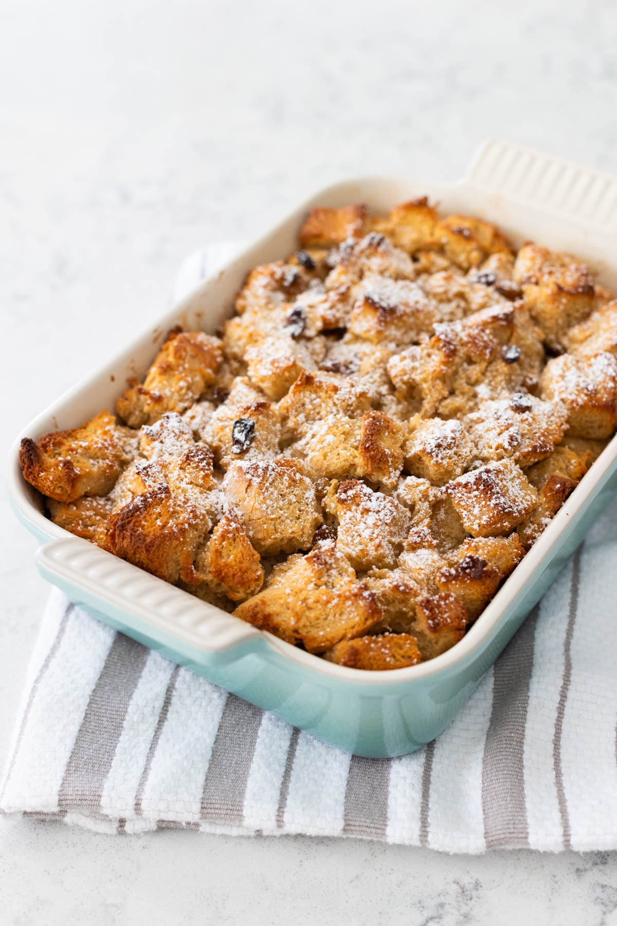 The baked bread pudding is golden brown. The dish sits on a towel as it cools. Powdered sugar has been sprinkled over the top for garnish.