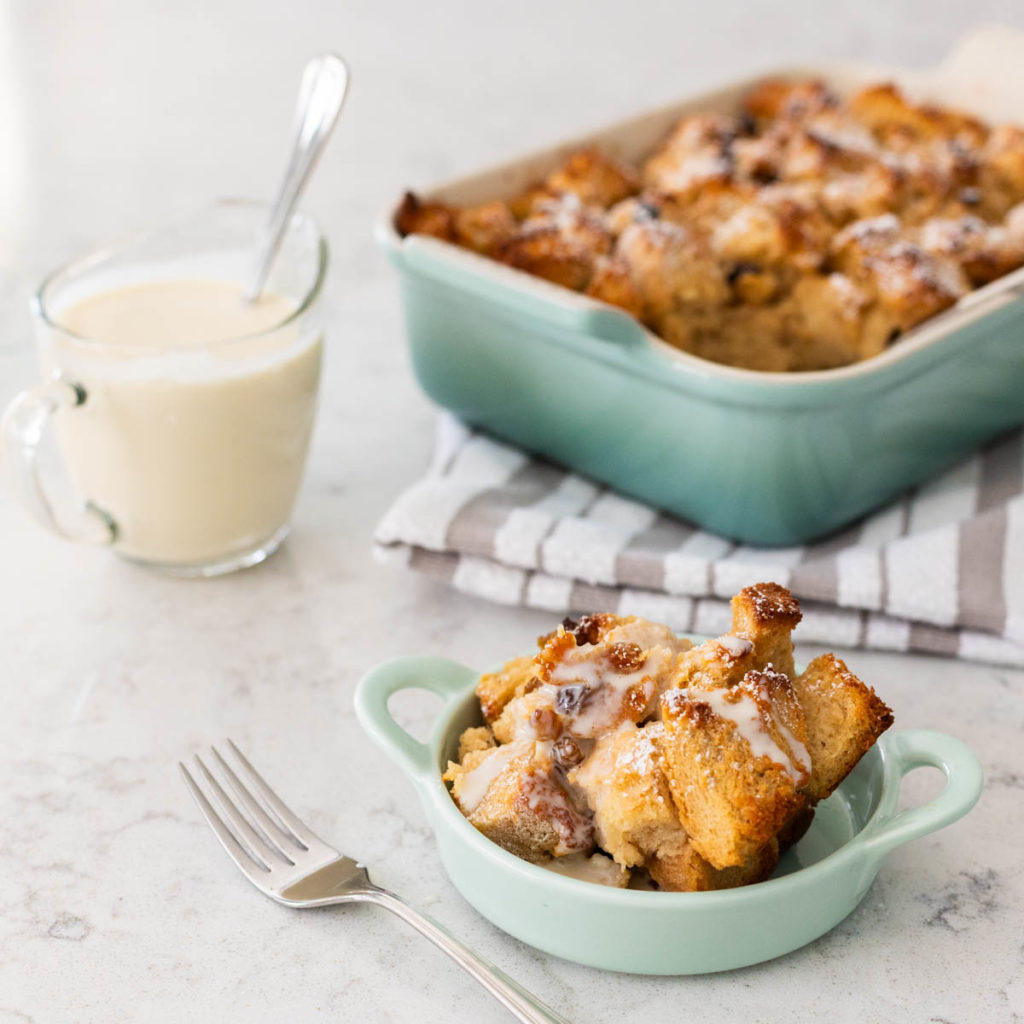 A baking pan of bread pudding sits next to a small dish with one serving that has a drizzle of the bourbon sauce over the top. The pitcher of bourbon sauce sits to the side.