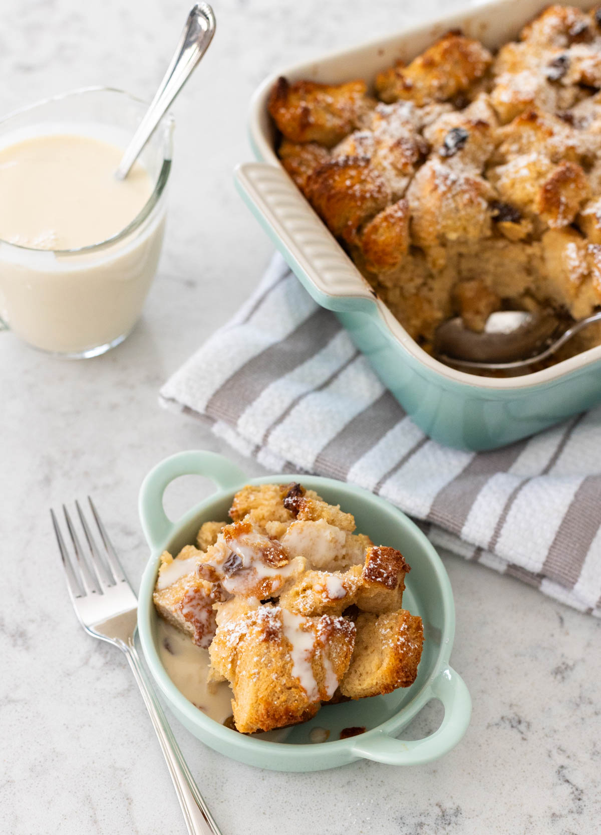 The bread pudding has been scooped into a small serving dish. A pitcher of bourbon sauce sits next to it and a little has been drizzled over the dessert.