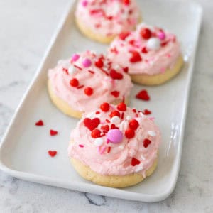 A white platter has pink frosted Valentine cookies with sweet red and pink sprinkles.