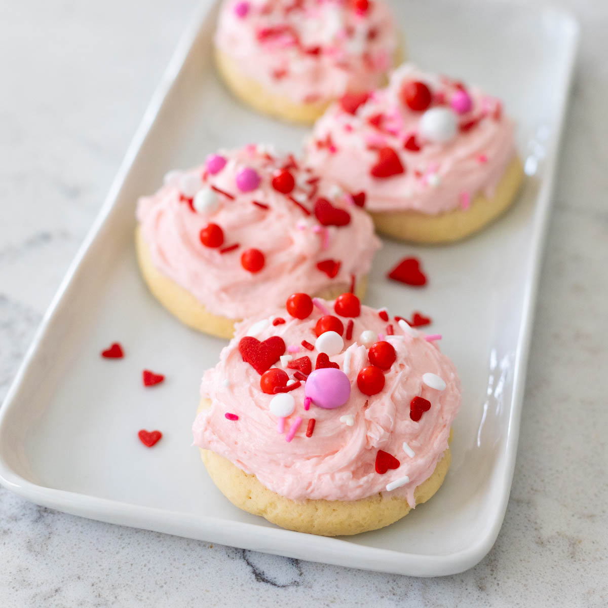 A white platter has pink frosted Valentine cookies with sweet red and pink sprinkles.