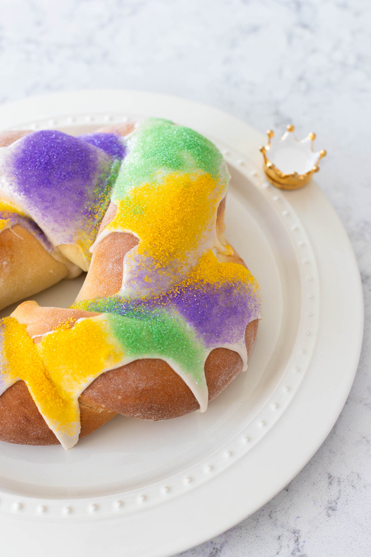 The finished king cake is on a platter with a crown decoration. The top is decorated with purple, green, and yellow sprinkles.