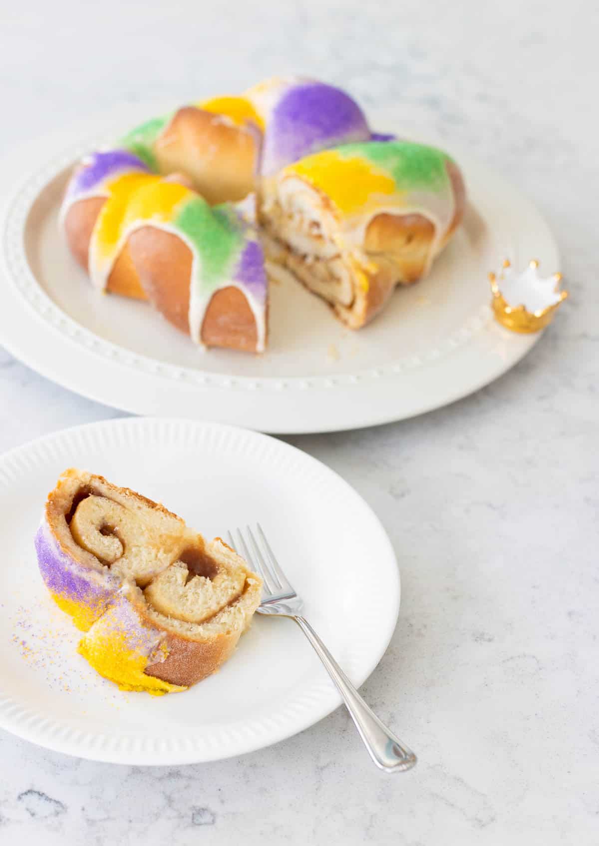 A slice of cinnamon king cake is on a plate next to the platter with the rest of the cake.