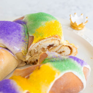 A braided King Cake has been sliced to show the cinnamon swirl filling. It is decorated with purple, green, and yellow sprinkles.