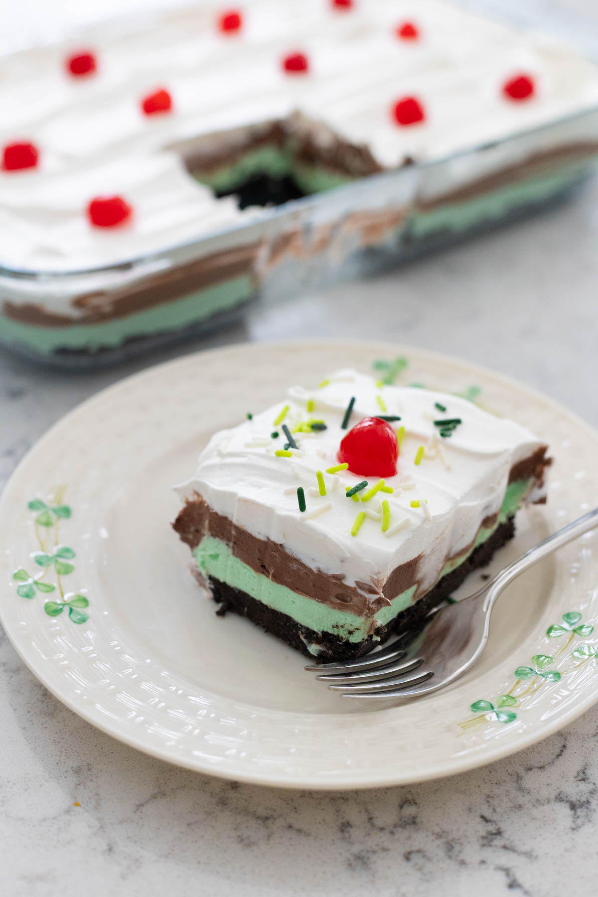 The layered no-bake dessert is on a party plate. You can see the light green cream cheese layer and chocolate pudding layer under the whipped topping and sprinkles.