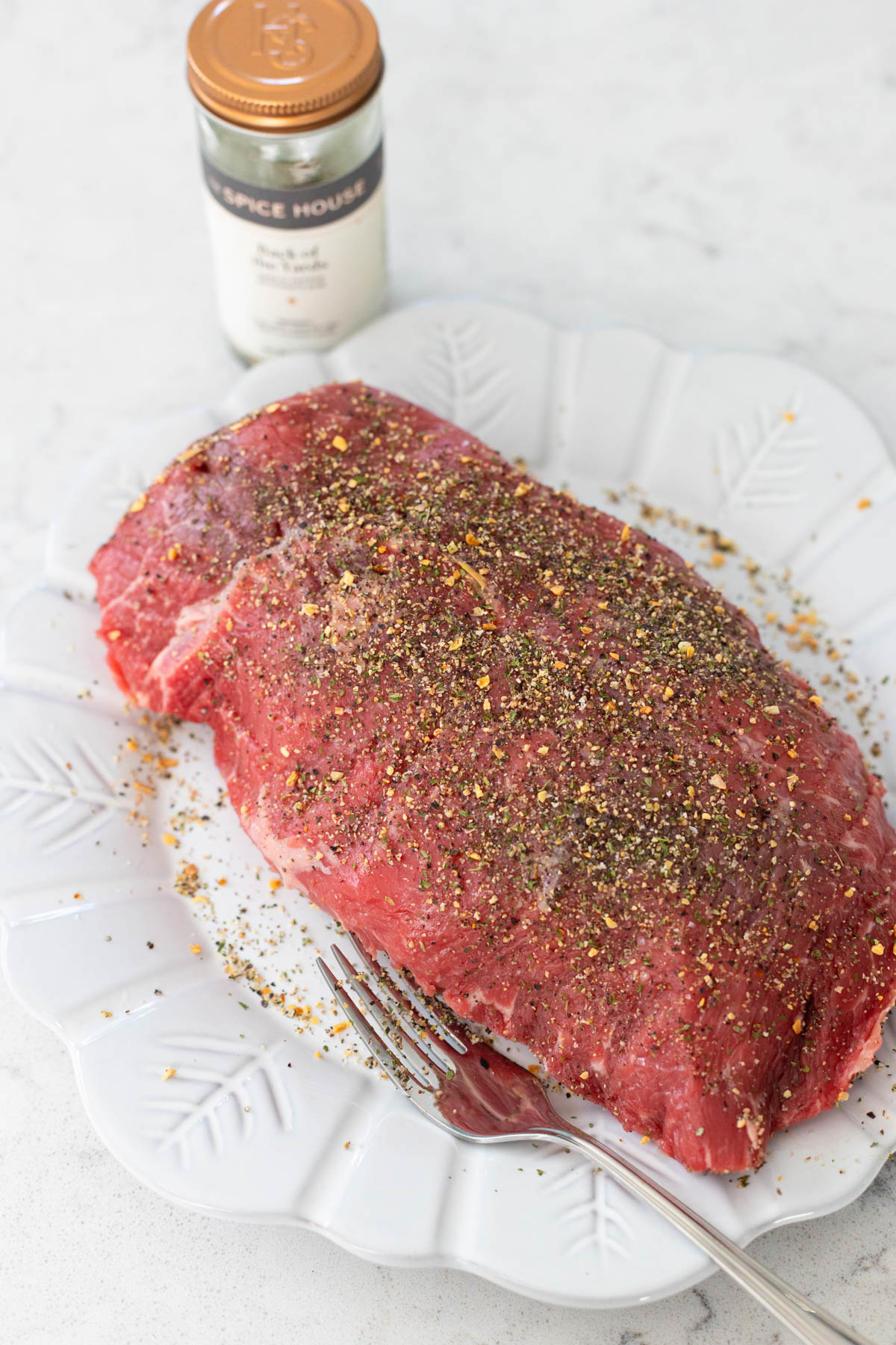 The chuck roast is on a dinner plate and has been sprinkled with seasonings on both sides.