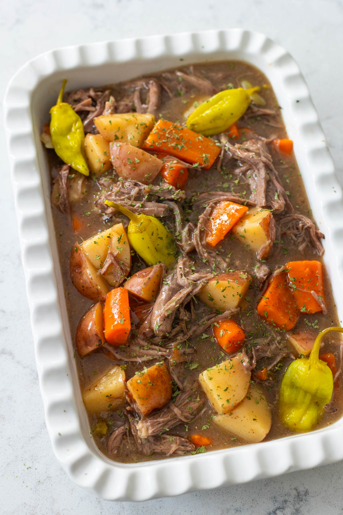 The finished pot roast mixture has been spooned into a baking dish for serving at the table. You can see large chunks of potatoes and carrots, pepperoncini peppers, and tender portions of shredded beef in a thick, rich gravy.