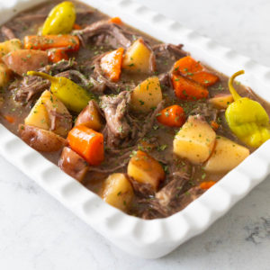 The white baking dish is filled with a chunky Mississippi pot roast with potatoes and carrots and pepperoncini peppers. There's a rich beefy gravy filling the dish.